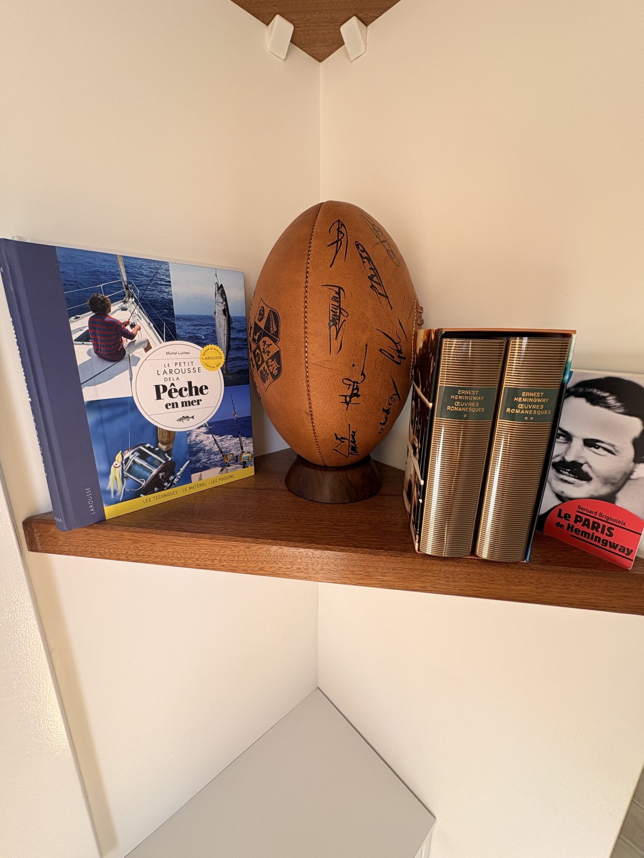 Uma estante com uma bola de futebol autografada, um livro intitulado 'Le Petit Larousse de la Pêche en mer' e dois livros de Ernest Hemingway.