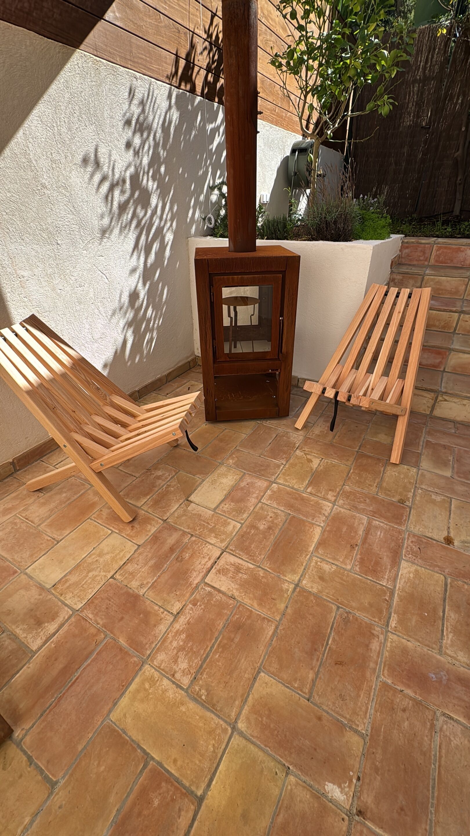 A patio area with a wood-burning stove, two wooden lounge chairs, and a stone-tiled floor.