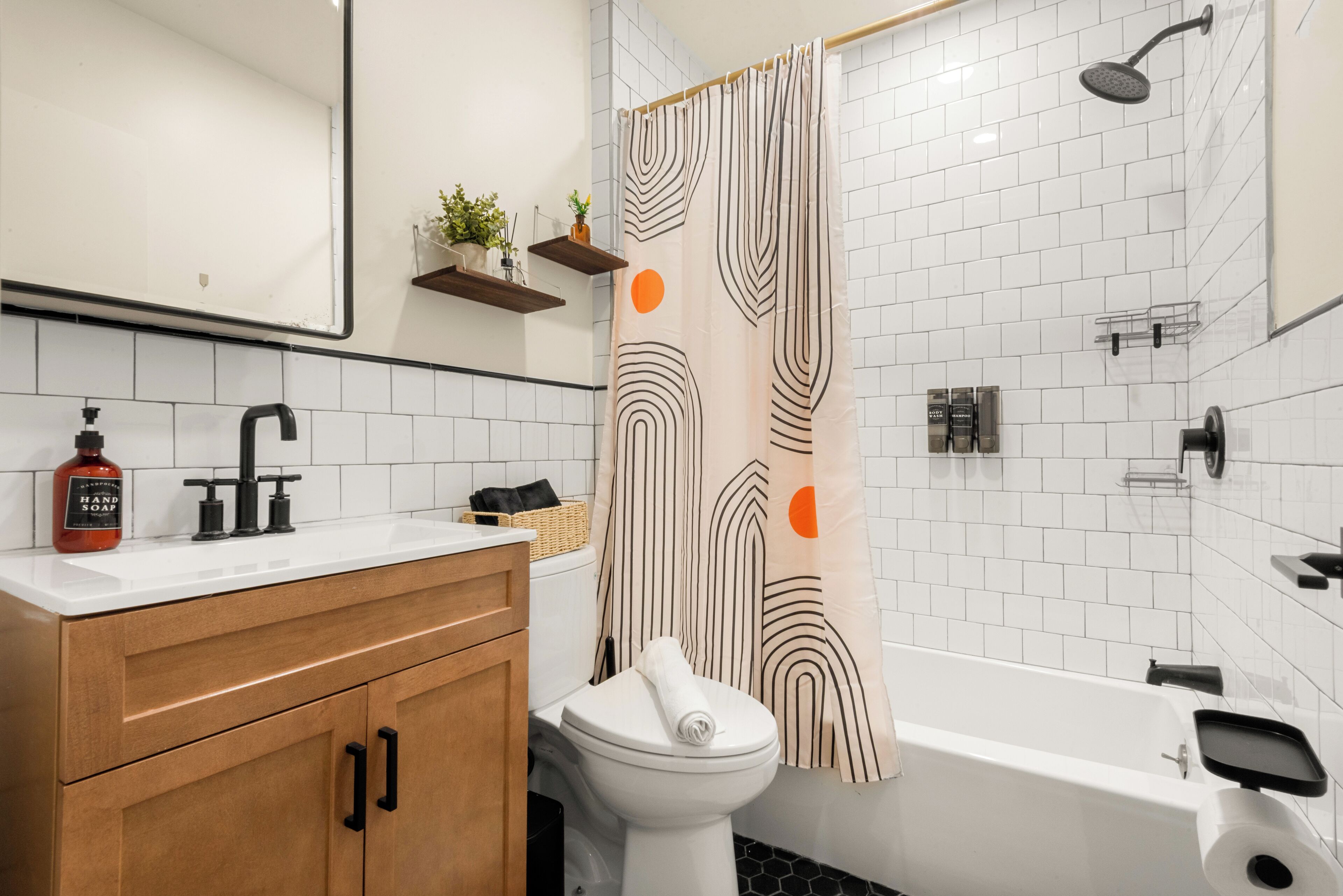 Et moderne badeværelse med en hvid flisemur, en trækommode, et hvidt toilet, en bruseniche med et håndholdt brusehoved i sort samt en mønstret bruseforhæng.