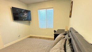 Een slaapkamer met een bed, een aan de muur bevestigde televisie, een raam met jaloezieën en een nachtkastje met een lamp.
