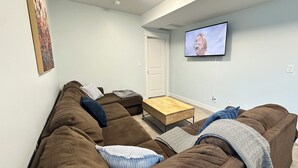 Een woonkamer met een bruine hoekbank, een houten salontafel en een flatscreen televisie aan de wand.