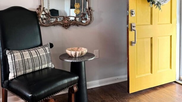Una habitación con un sillón de cuero negro, una mesa redonda con un cuenco, un espejo con un marco decorativo y una puerta amarilla con un adorno.