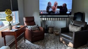 Una sala de estar con un televisor de pantalla plana montado en la pared, un ventilador de techo y dos sillones de cuero.