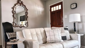 Un salón con un sofá de cuero blanco, una mesa de centro redonda, una silla negra, un espejo decorativo, un reloj y una lámpara.