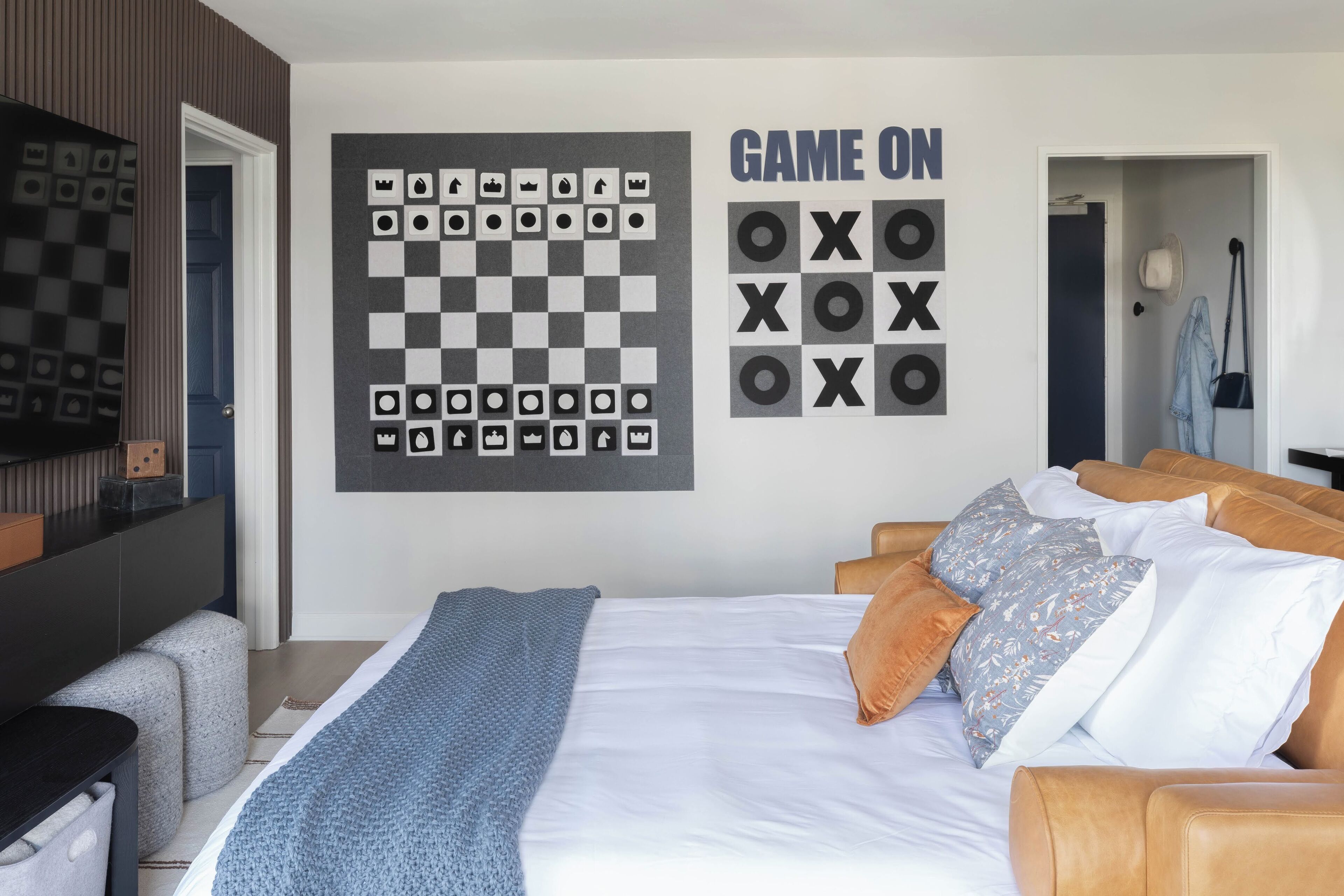 A bedroom with a bed, pillows, a blanket, a wall-mounted chessboard, and a sign that says "GAME ON".
