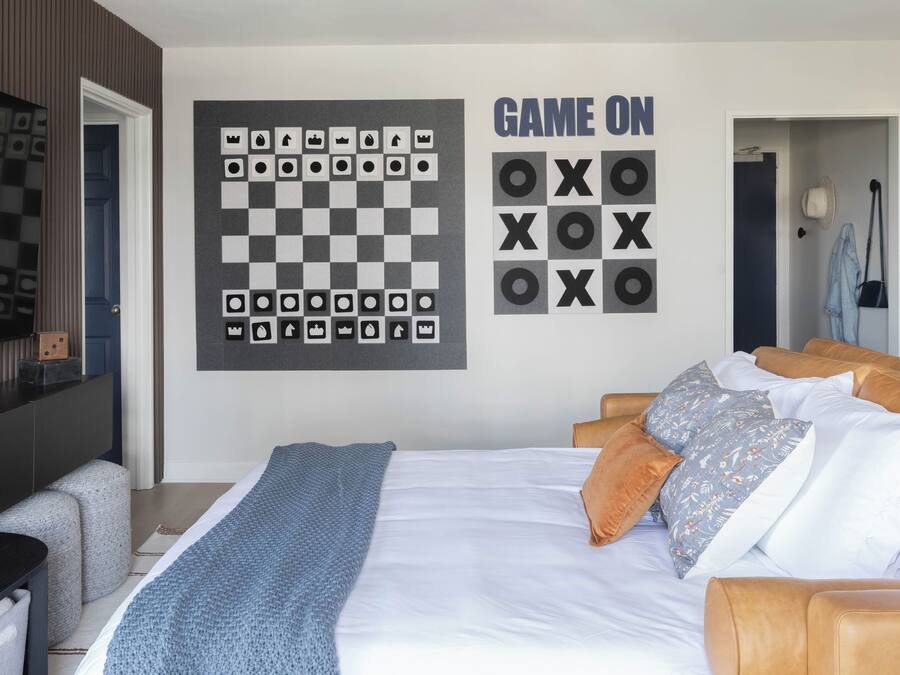 A bedroom with a bed, pillows, a blanket, a wall-mounted chessboard, and a sign that says "GAME ON".