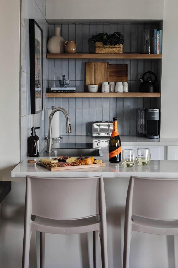 A modern kitchen with a white countertop, two bar stools, a wine bottle, and a variety of kitchen appliances. - Downtown ATL Condo Balcony (Atlanta)