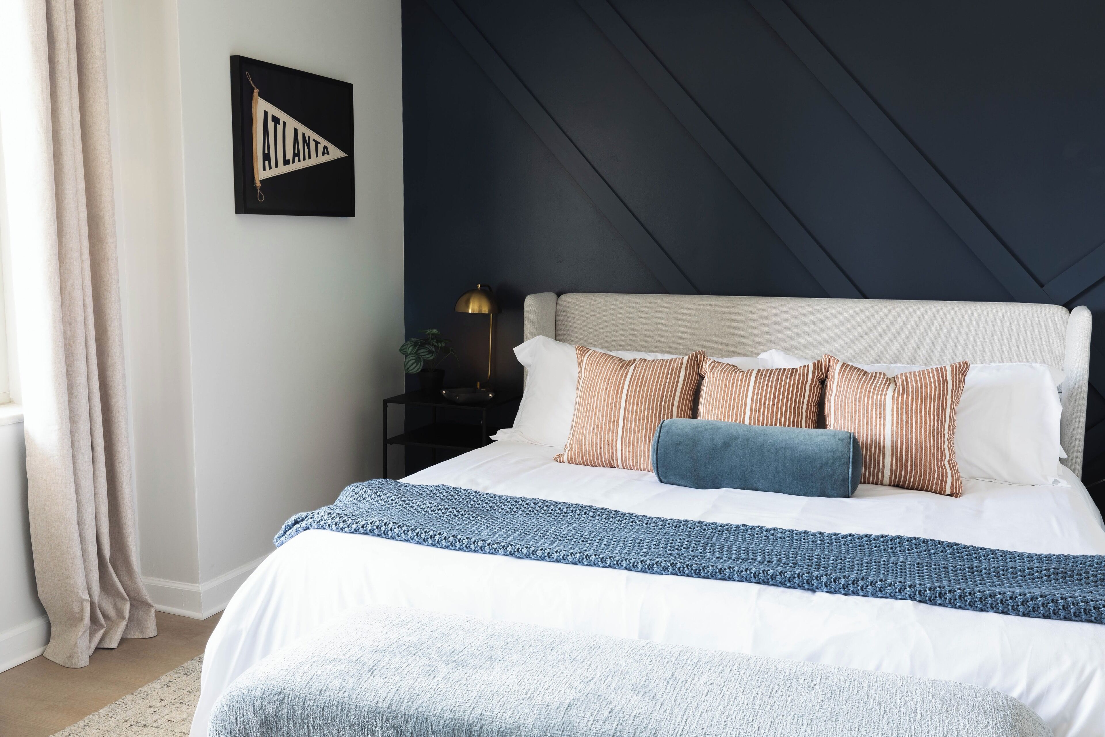 Un lit bien fait, recouvert d’une couverture blanche et bleue, un mur foncé orné d’un motif géométrique et un panneau encadré accroché au mur.