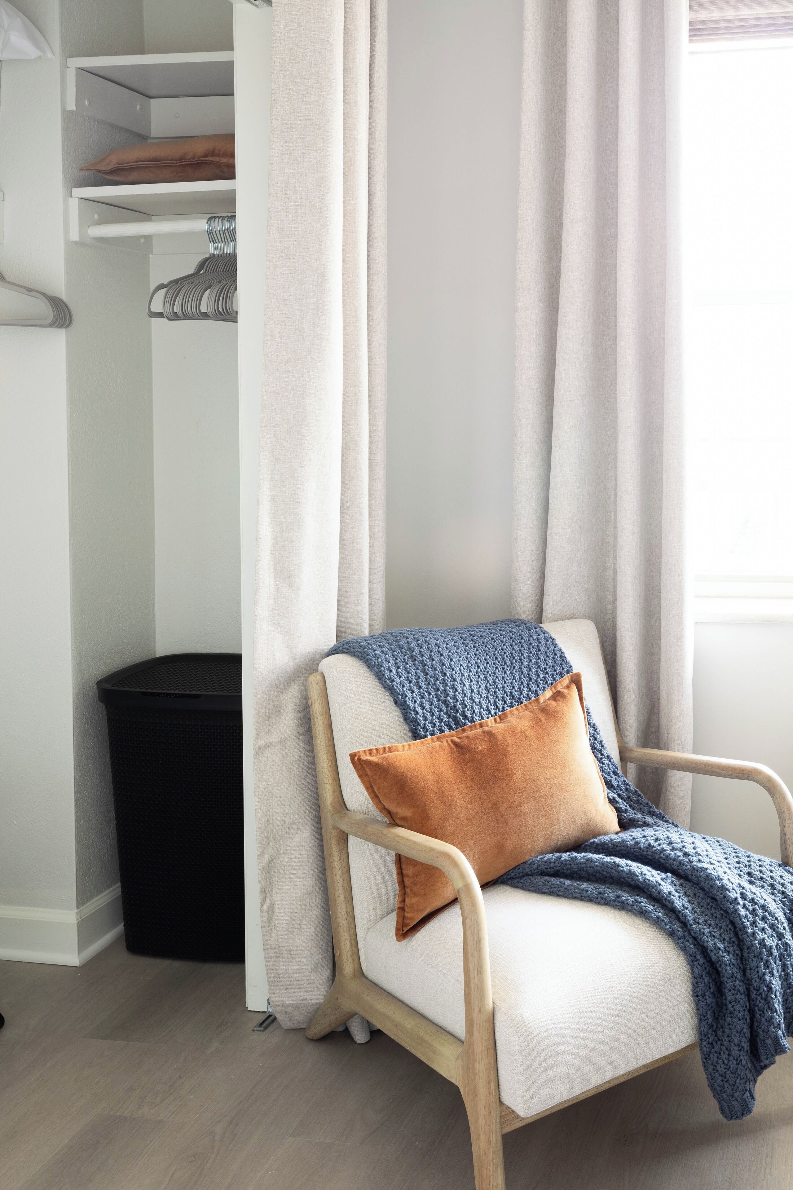 Una silla con un cojín blanco y una manta tejida azul, una almohada marrón, y un bote de basura negro en una habitación con una pared blanca y una ventana con cortinas de tul.