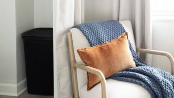 A chair with a white cushion and a blue knitted throw, a brown pillow, and a black trash bin in a room with a white wall and a window with sheer curtains.
