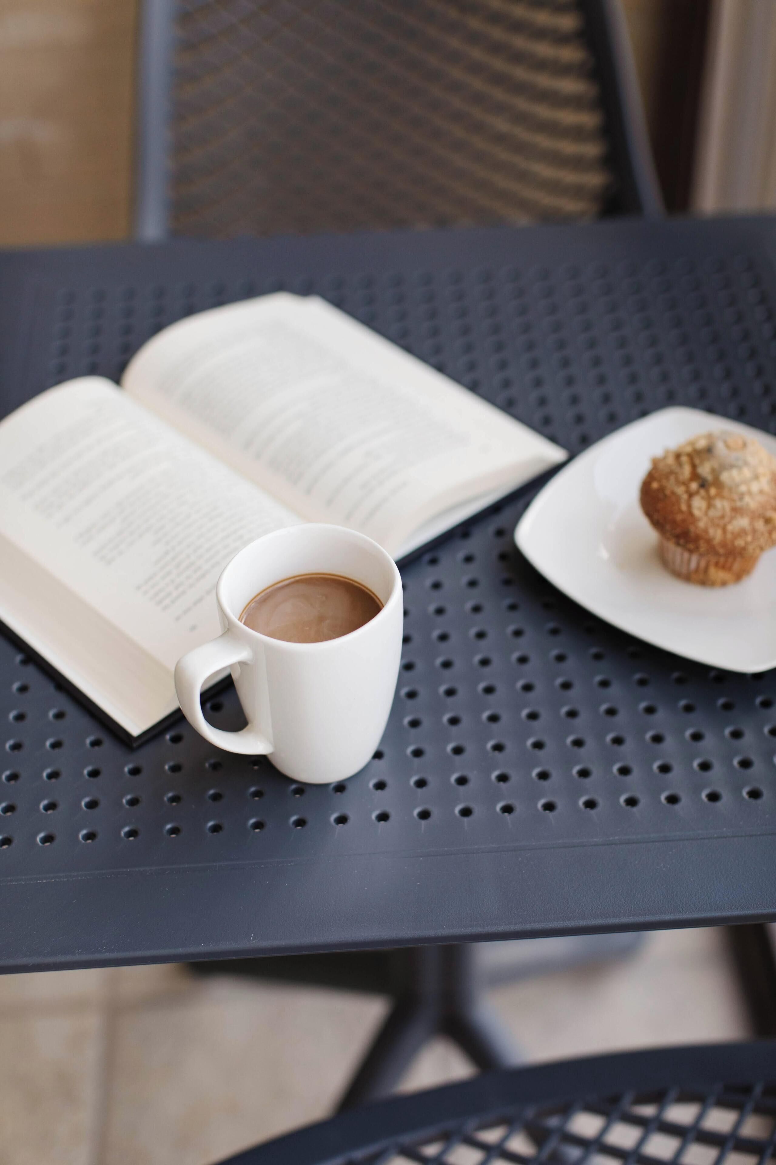 An open book, a white coffee cup, and a muffin on a perforated metal table.