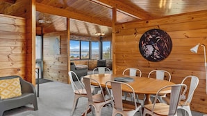 A wooden cabin interior with a dining area, a sofa with a patterned cushion, and a large window offering a scenic view.