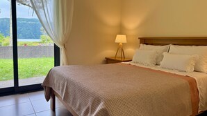 A hotel room with a large bed, a bedside table with a lamp, and a view of greenery and a body of water through a window. - LAKEVIEW GREEN PARADISE By Azores HomeMade (Ponta Delgada)