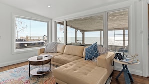 A modern living room with a large sectional sofa, a small round table, and a view of the ocean through large windows. - Lakeview Escape. Hot Tub, Sauna, Screened Porch. Birding Paradise. Wineries! (Amherstburg)