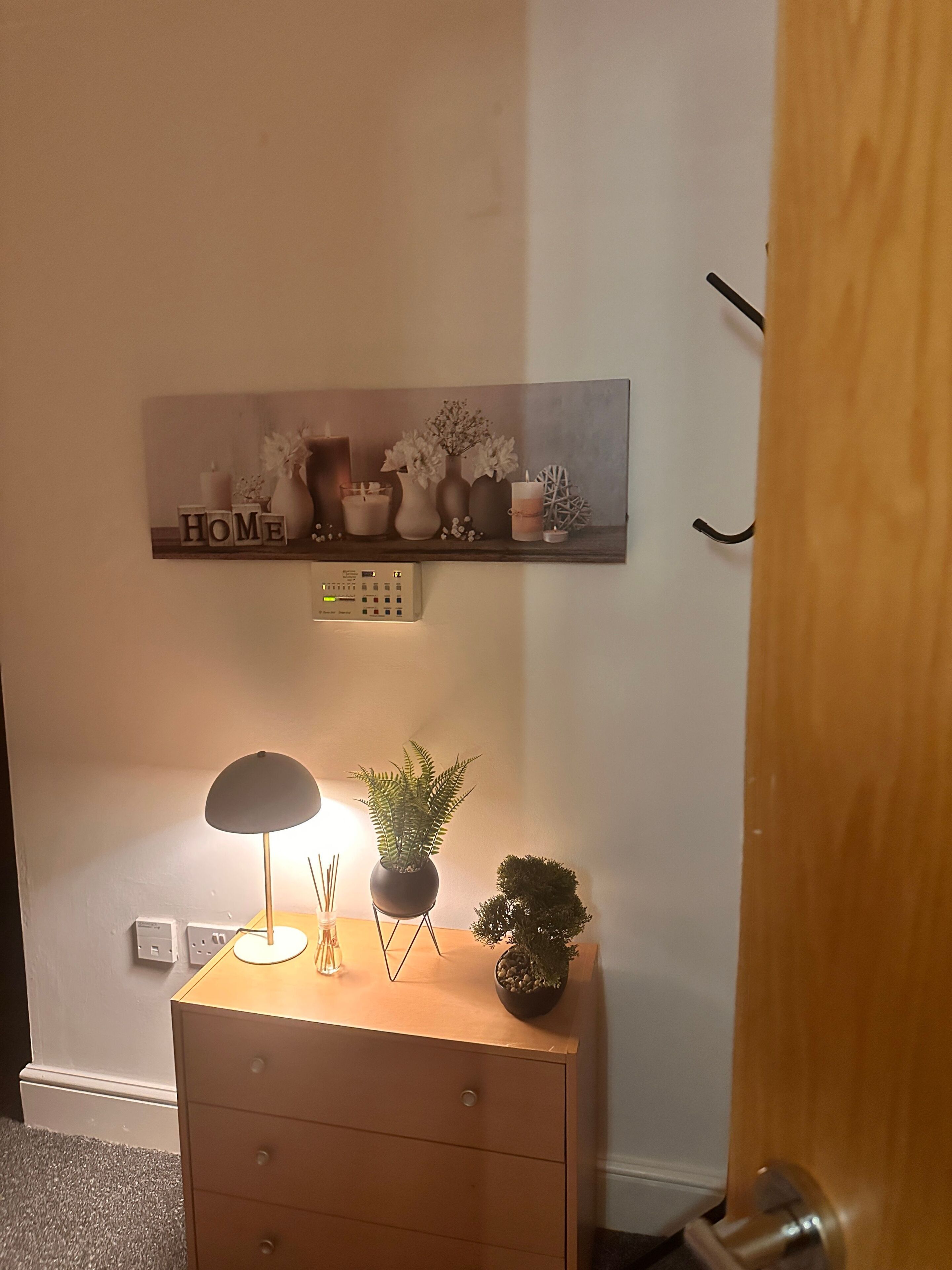 A room with a wooden dresser, a wall-mounted shelf with decorative items, a lamp, and a small potted plant.