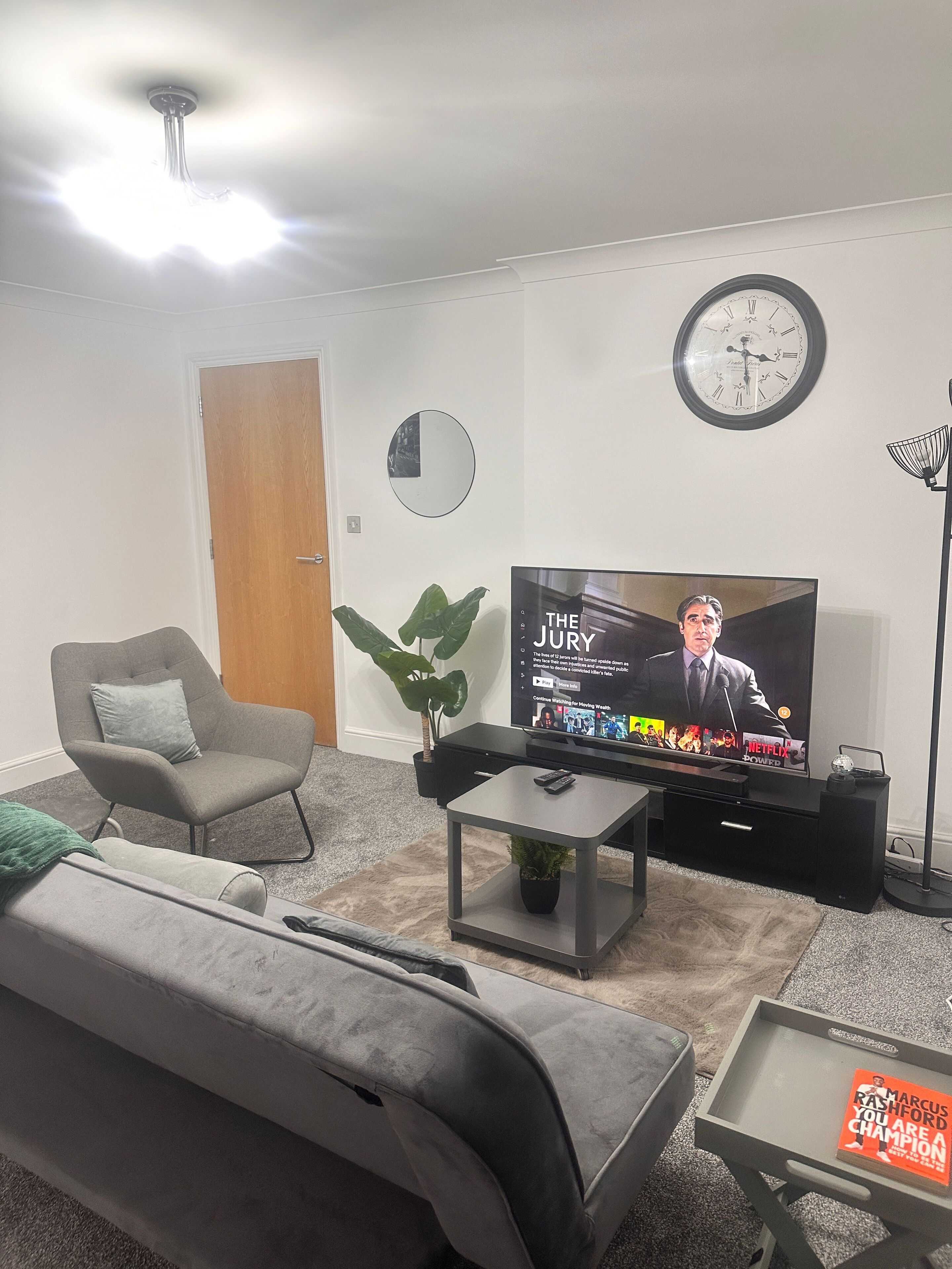 Una sala de estar con un sofá gris, una mesa de centro con una planta, un televisor mostrando un noticiero, un reloj y una puerta.