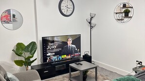 A modern living room with a flat-screen TV, a sofa, a coffee table, a floor lamp, and a wall clock. - One Bedoom Flat in Birmingham City Centre With Free Underground Parking (Birmingham)