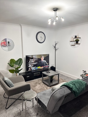A modern living room with a flat-screen TV, a sofa, a coffee table, a side table, a potted plant, a wall clock, and a decorative light fixture. - One Bedoom Flat in Birmingham City Centre With Free Underground Parking (Birmingham)