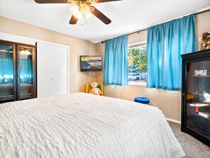 Un dormitorio con cama, ventilador de techo, ventana con cortinas, televisor y armario.