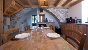 A dining area with a long table set for six, stone walls, wooden beams, and a kitchen in the background.