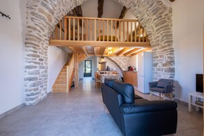 A spacious living area with a stone wall, a wooden staircase, a black leather sofa, and a kitchen in the background. - Le P'tit Pelet - FRAISSINET-DE-FOURQUES (FRAISSINET-DE-FOURQUES)