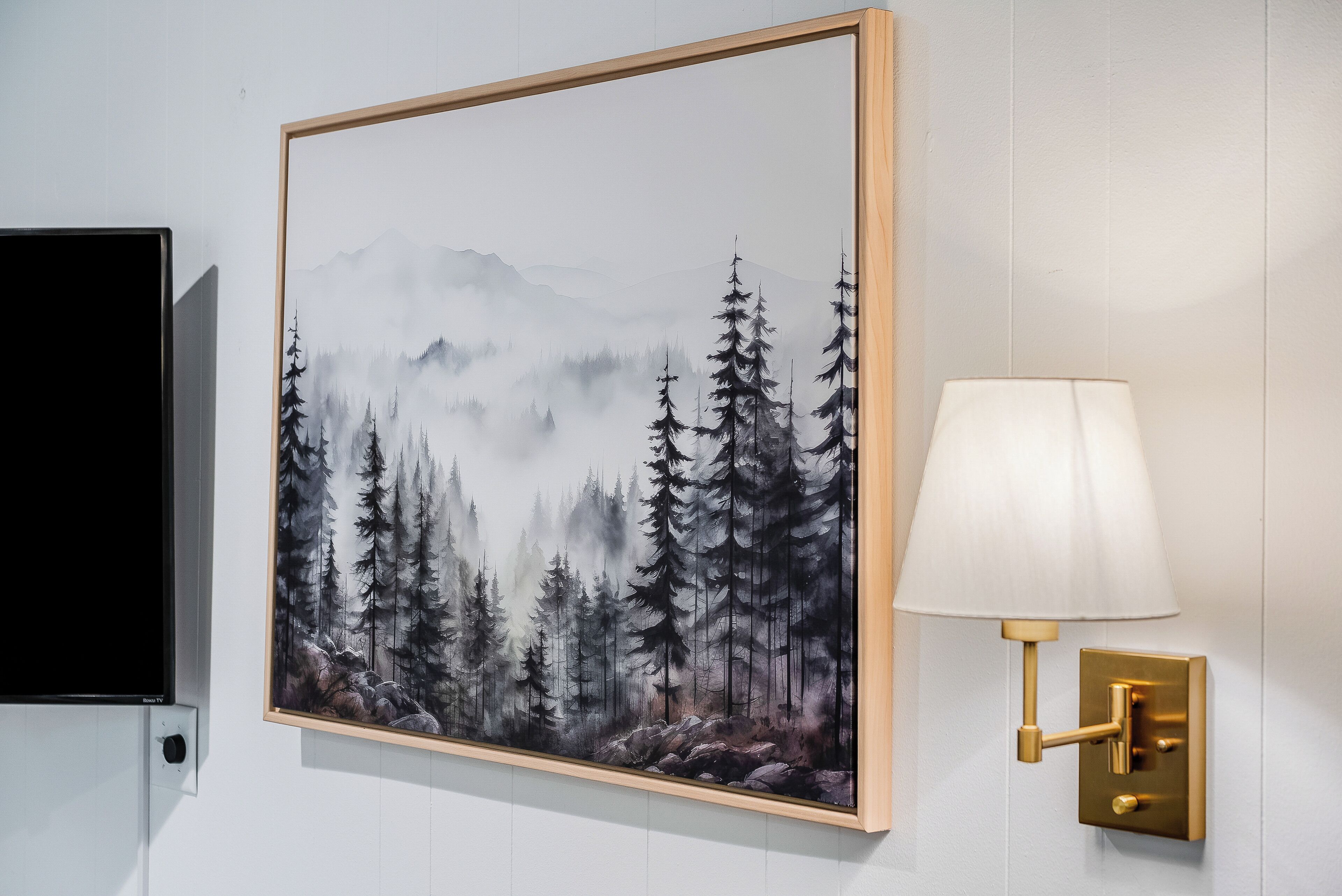Ein gerahmtes Bild von einem Wald mit Bergen, eine Wandleuchte und ein Fernseher an der Wand.