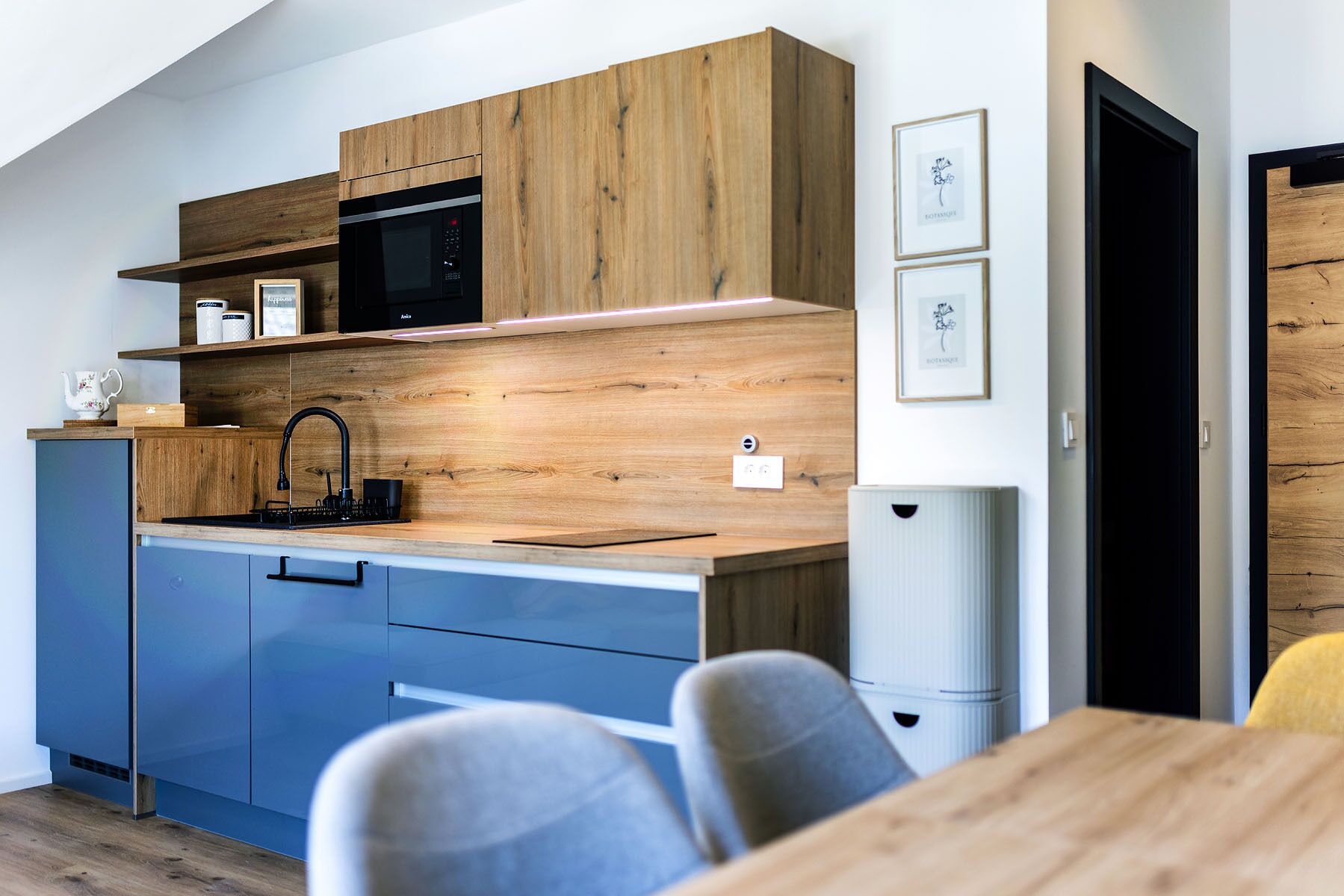 A modern kitchen with wooden cabinetry, a black microwave, and a sink. There are framed pictures on the wall and a door to the right.