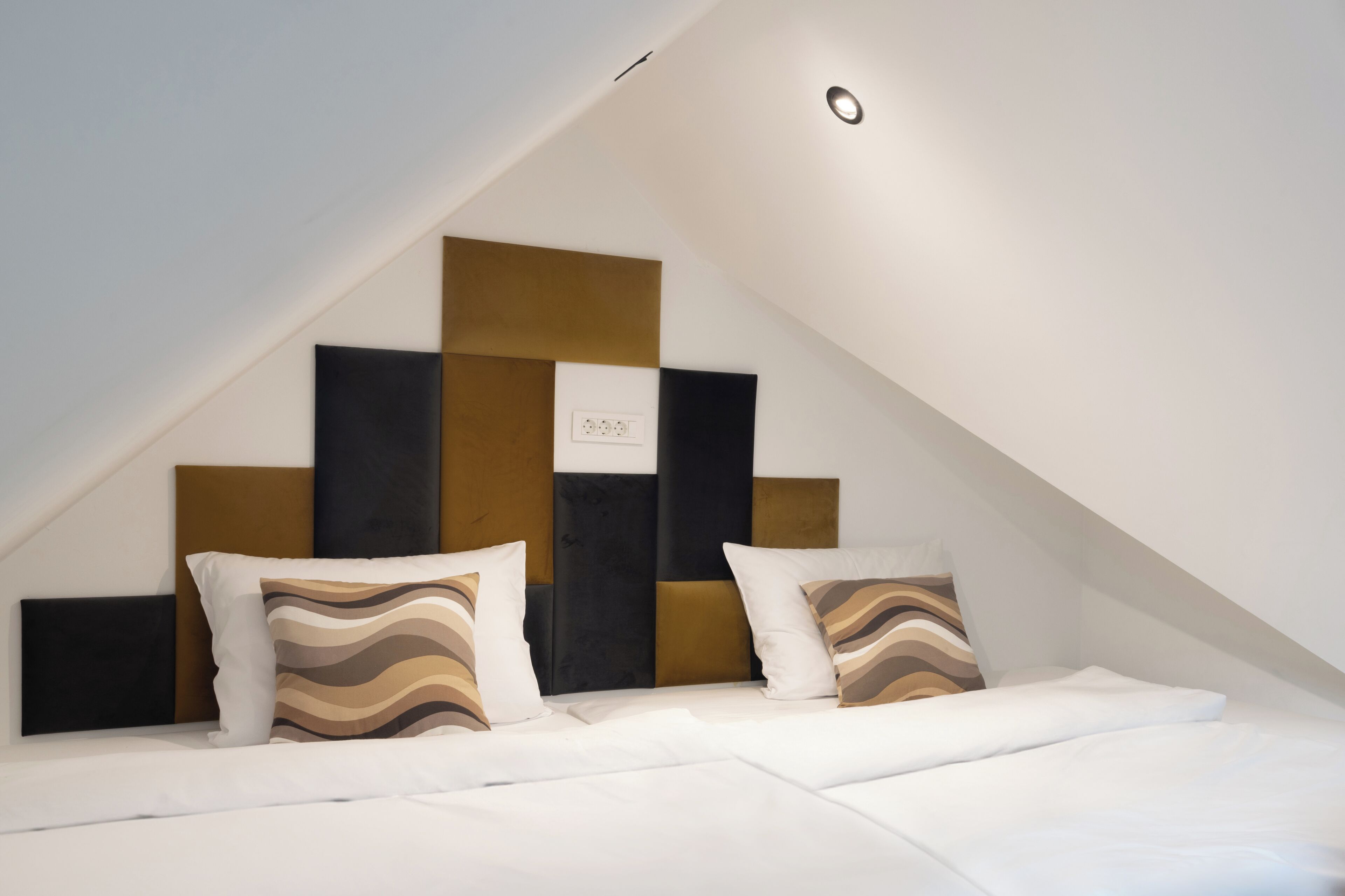 Une chambre d’hôtel moderne avec un plafond en pente, un lit recouvert de draps blancs et une tête de lit ornée de motifs géométriques.