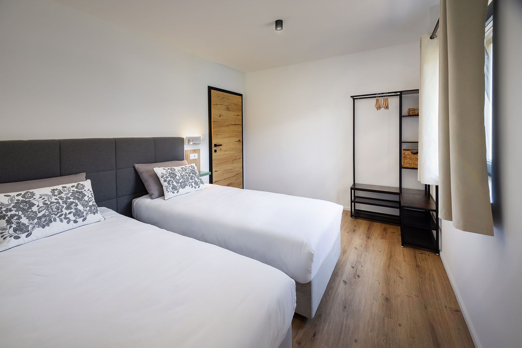 A hotel room with two beds, a wooden door, a small desk with shelves, and a window with curtains.