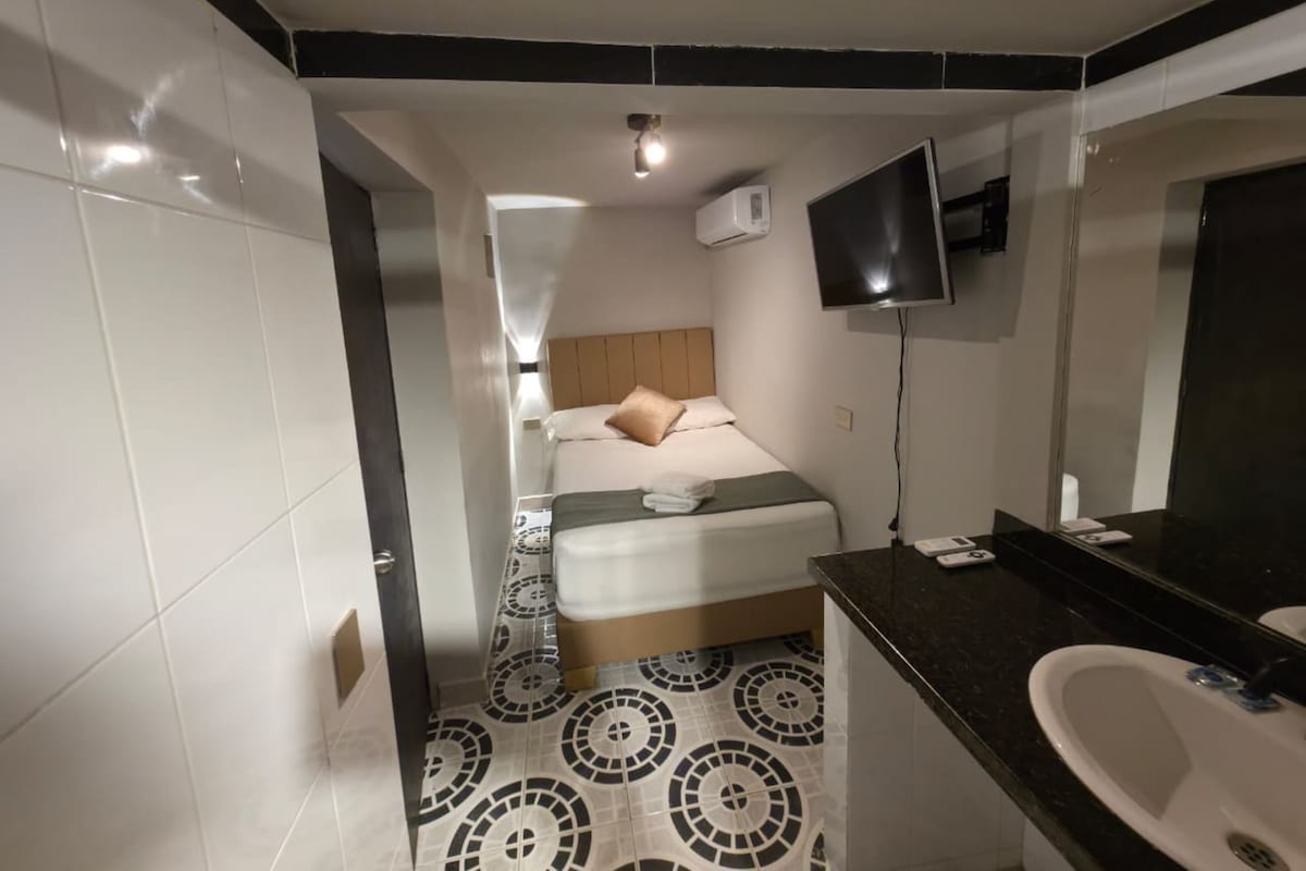 Une chambre d’hôtel avec un lit, un téléviseur fixé au mur, une salle de bain avec un lavabo et un miroir, et un sol à motifs.