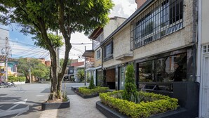 Vista de la calle con un edificio que tiene grandes ventanales y un balcón. Hay macetas con plantas frente al edificio.