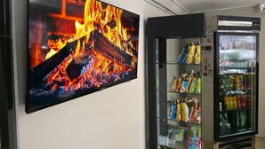 Un televisor de pantalla plana montado en la pared mostrando un fuego, una máquina expendedora con aperitivos y un refrigerador lleno de bebidas.