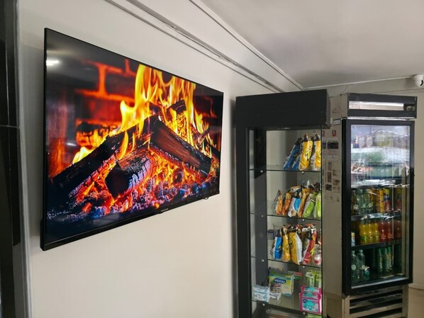 Televisão de tela plana presa na parede exibindo uma lareira, uma máquina de venda automática com lanches e uma geladeira abastecida com bebidas.