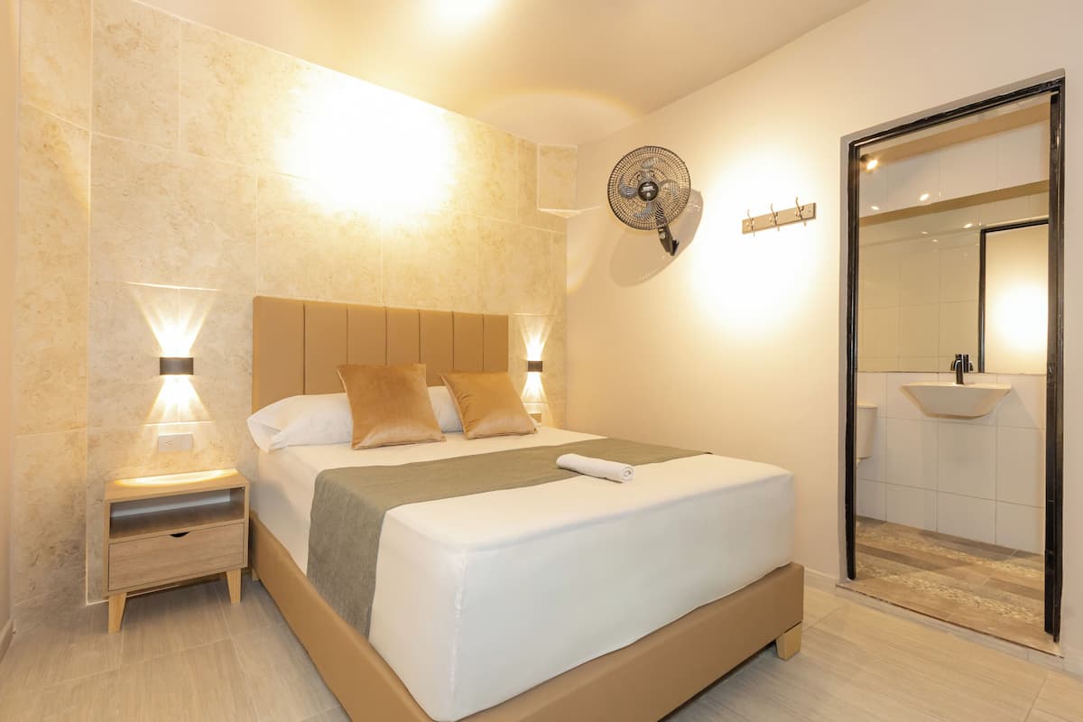 Moderni hotellihuone, jossa on suuri sänky, yöpöytävalaisimet, seinään kiinnitetty tuuletin sekä kylpyhuone, jossa on pesuallas ja peili.