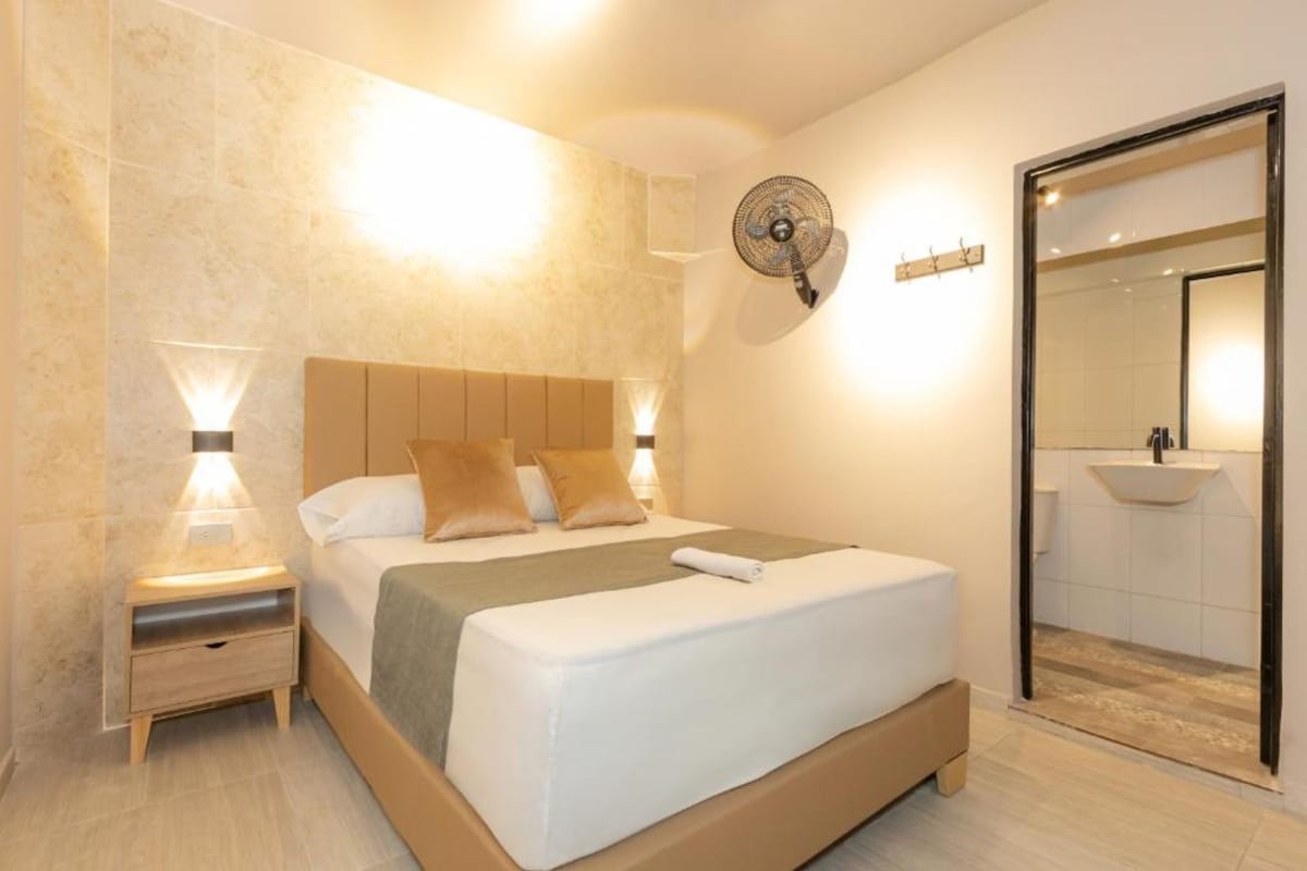 Une chambre moderne avec un grand lit, des lampes de chevet, un ventilateur fixé au mur et une salle de bain équipée d’un lavabo et d’un miroir.