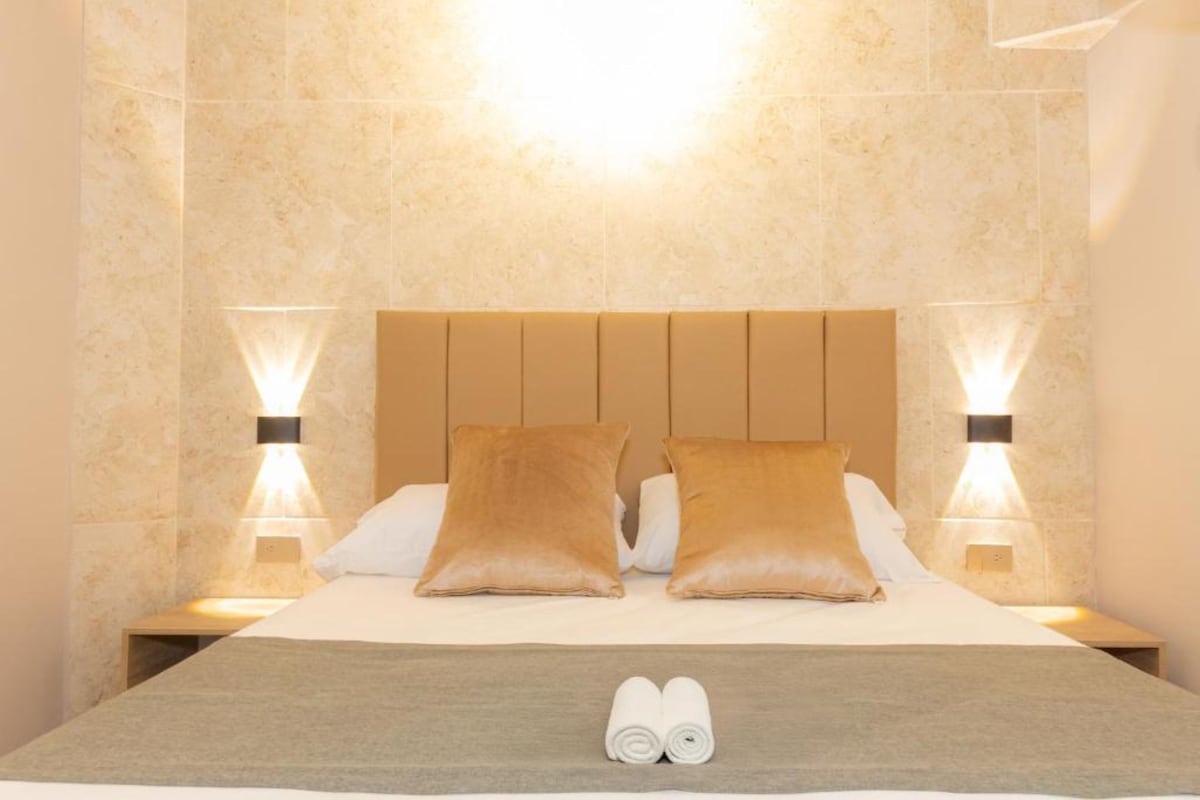 Una habitación de hotel con una cama, dos mesitas de noche, dos lámparas de pared y un par de toallas enrolladas sobre la cama.