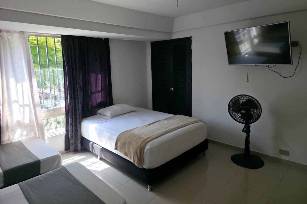 Une chambre avec un lit, une télévision, un ventilateur et une vue sur la verdure environnante.