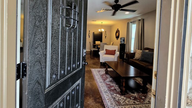 Una sala de estar con una puerta de madera oscura, un ventilador de techo, un comedor con mesa y sillas, y un sofá.