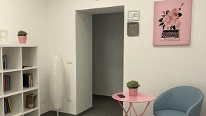 Una habitación moderna con una estantería, una pequeña mesa rosa con una planta, una silla azul, un reloj y un equipo de aire acondicionado.
