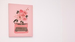 Una habitación con un sillón azul claro, una mesa rosa con una planta y un teléfono inteligente, y un cuadro con marco rosa en la pared.