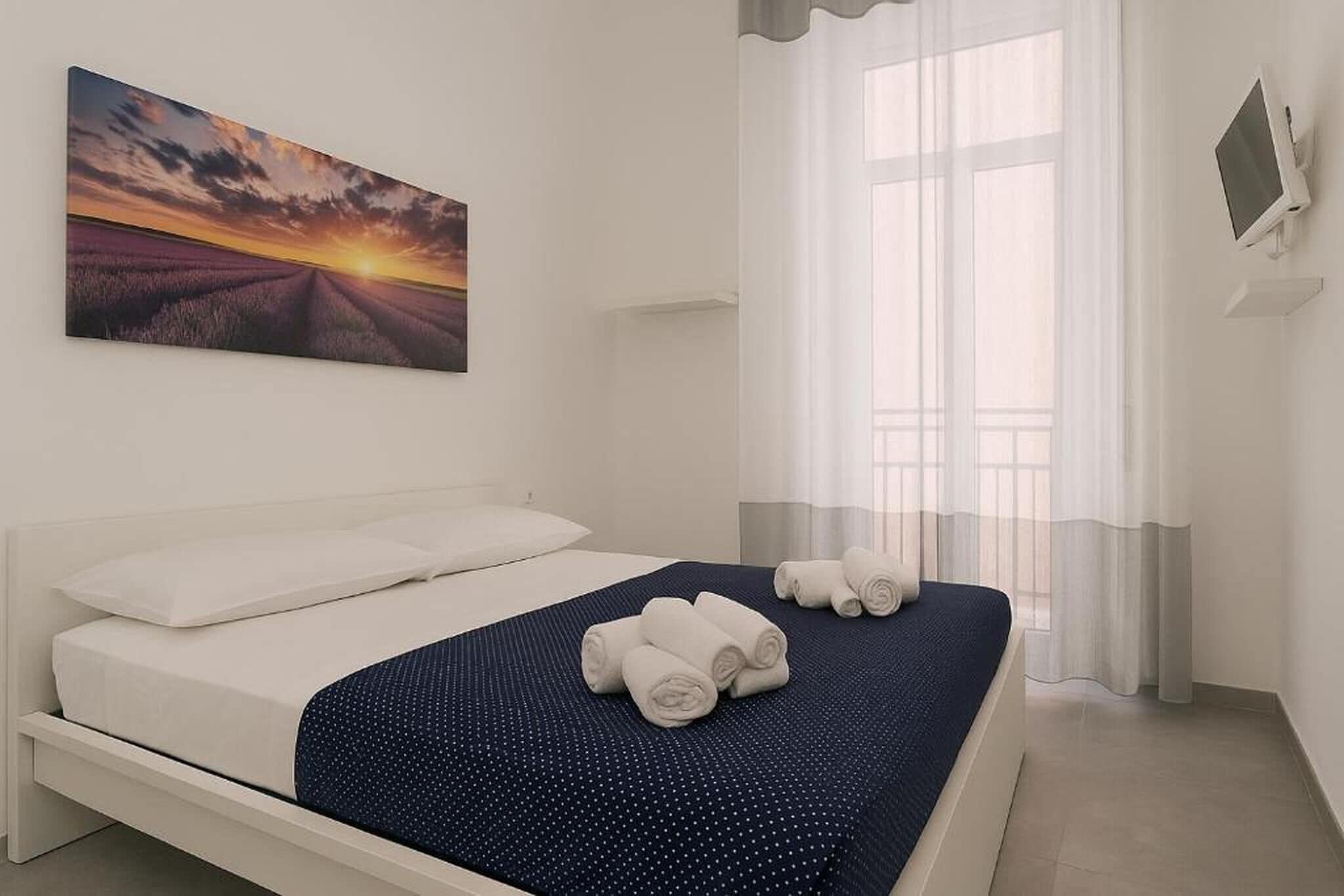 Ein ordentlich bezogenes Bett mit weißen Laken und einer Bettdecke mit blauen Punkten, ein aufgerolltes weißes Handtuch auf dem Bett und ein Sonnenuntergangsbild an der Wand.