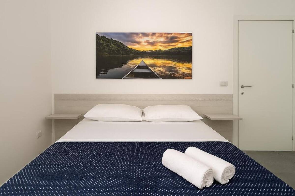 Hotellihuone, jossa on sänky, kaksi tyynyä, sinivalkoinen pilkkukuviollinen päiväpeite ja maalaus, jossa on kuvattu auringonlasku joen yllä.