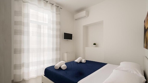 Una habitación de hotel con cama, televisión, una planta pequeña y una ventana con cortina translúcida.