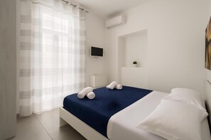Une chambre d’hôtel avec un lit, une télévision, une petite plante et une fenêtre avec un rideau léger.