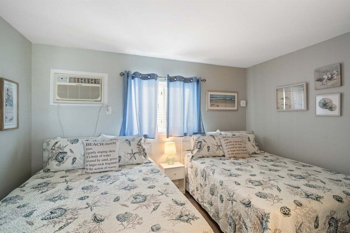 Ein Zimmer mit zwei Betten, einem fensterbelaubten Fenster, Wandklimaanlage und gerahmten Bildern.