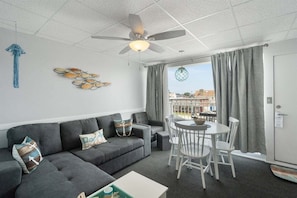 Ein modernes Wohnzimmer mit einem grauen Sofa, einem Esstisch mit Stühlen, einem Deckenventilator und Blick durch große Fenster.