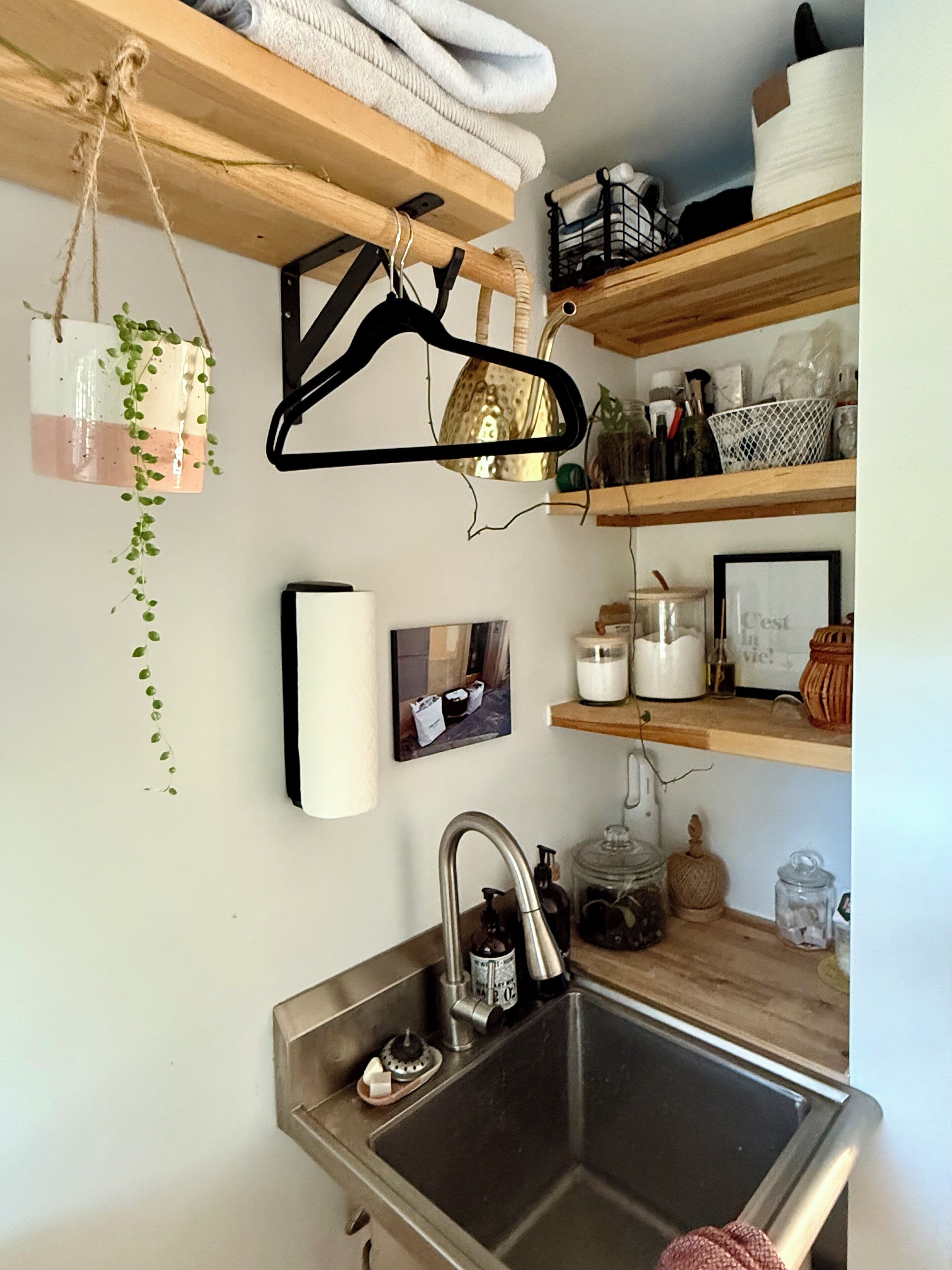 A kitchen with a stainless steel sink, wooden shelves, and hanging items like a plant and a black hanger.