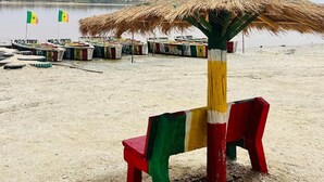 A beach with a thatched umbrella and colorful chairs, a sandy shore, and flags in the background. - Authentic stay at Villa Noflaye du Lac Rose! (Deni Biram Ndao)
