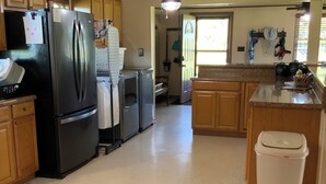 Una cocina con armarios de madera, un refrigerador negro, un basurero blanco y una ventana con persianas.