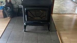 Un poêle à bois, avec un mur en pierre en arrière-plan, un sol carrelé et un coin cuisine à l’arrière-plan.